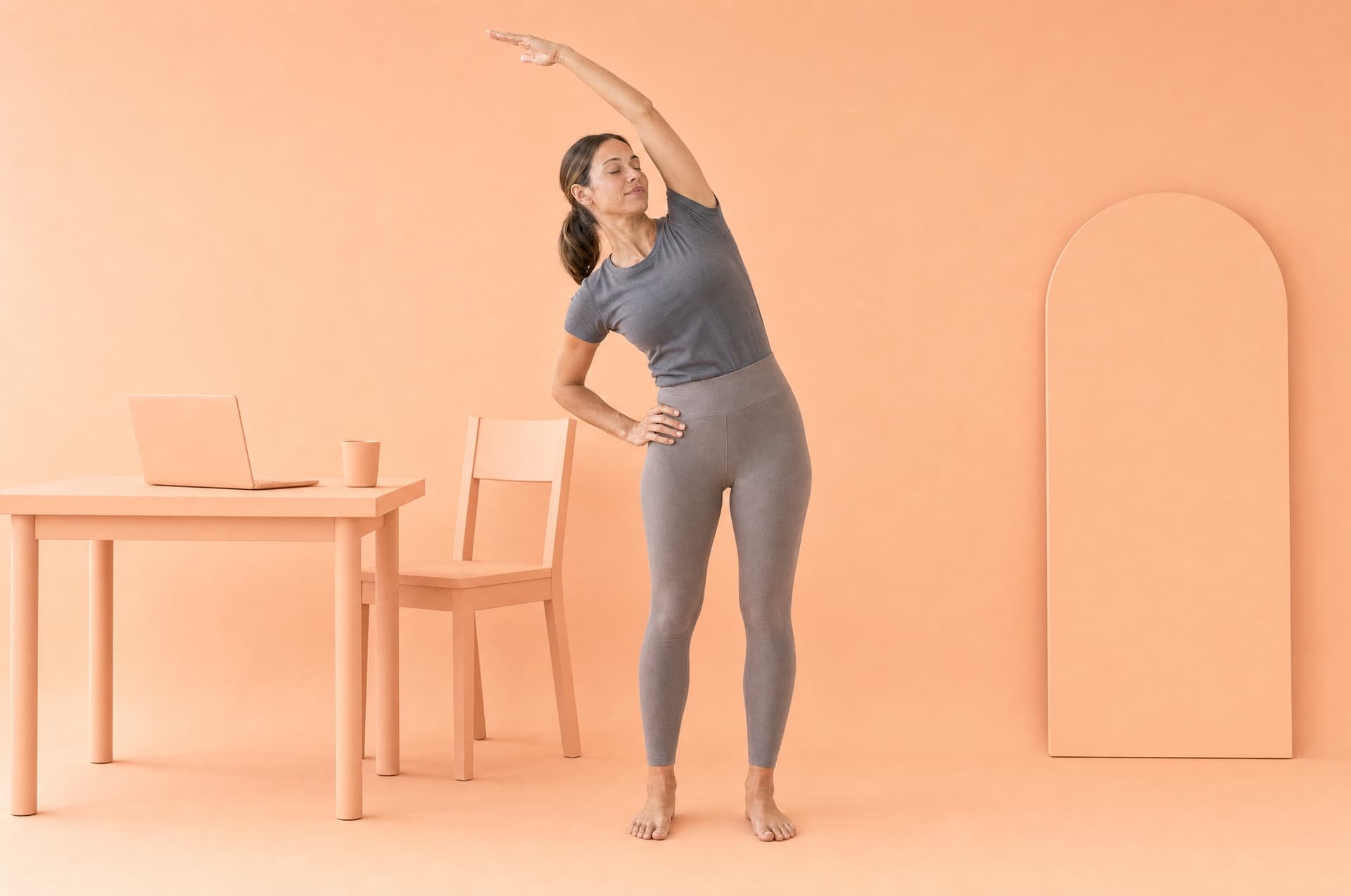 A woman standing in a peach studio in a gentle side bend, one arm overhead, an open laptop on the desk behind her.