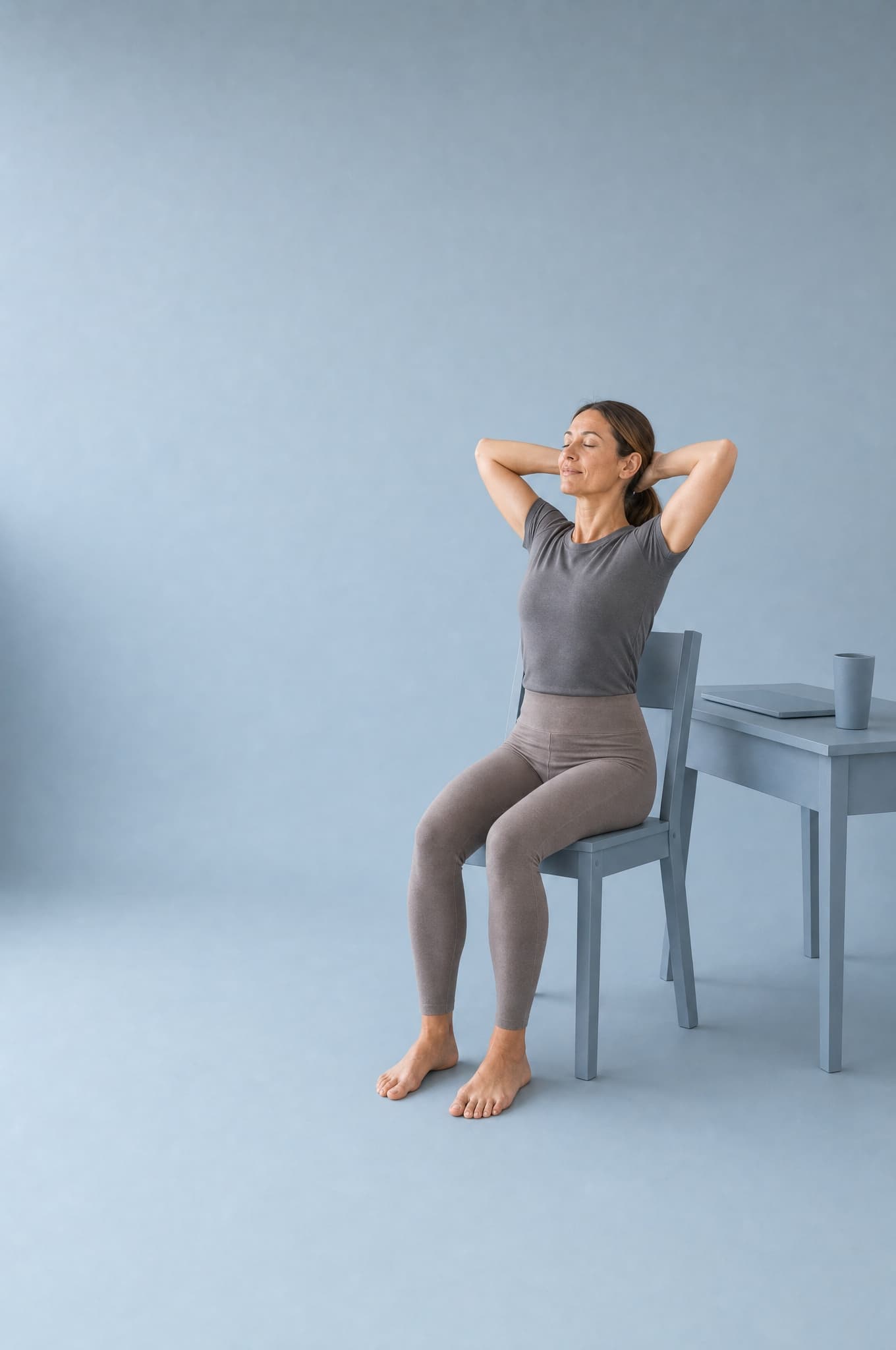 A woman seated on a slate-blue chair in a slate-blue studio, hands behind her head, elbows open, eyes closed, a closed laptop on a small desk behind her.