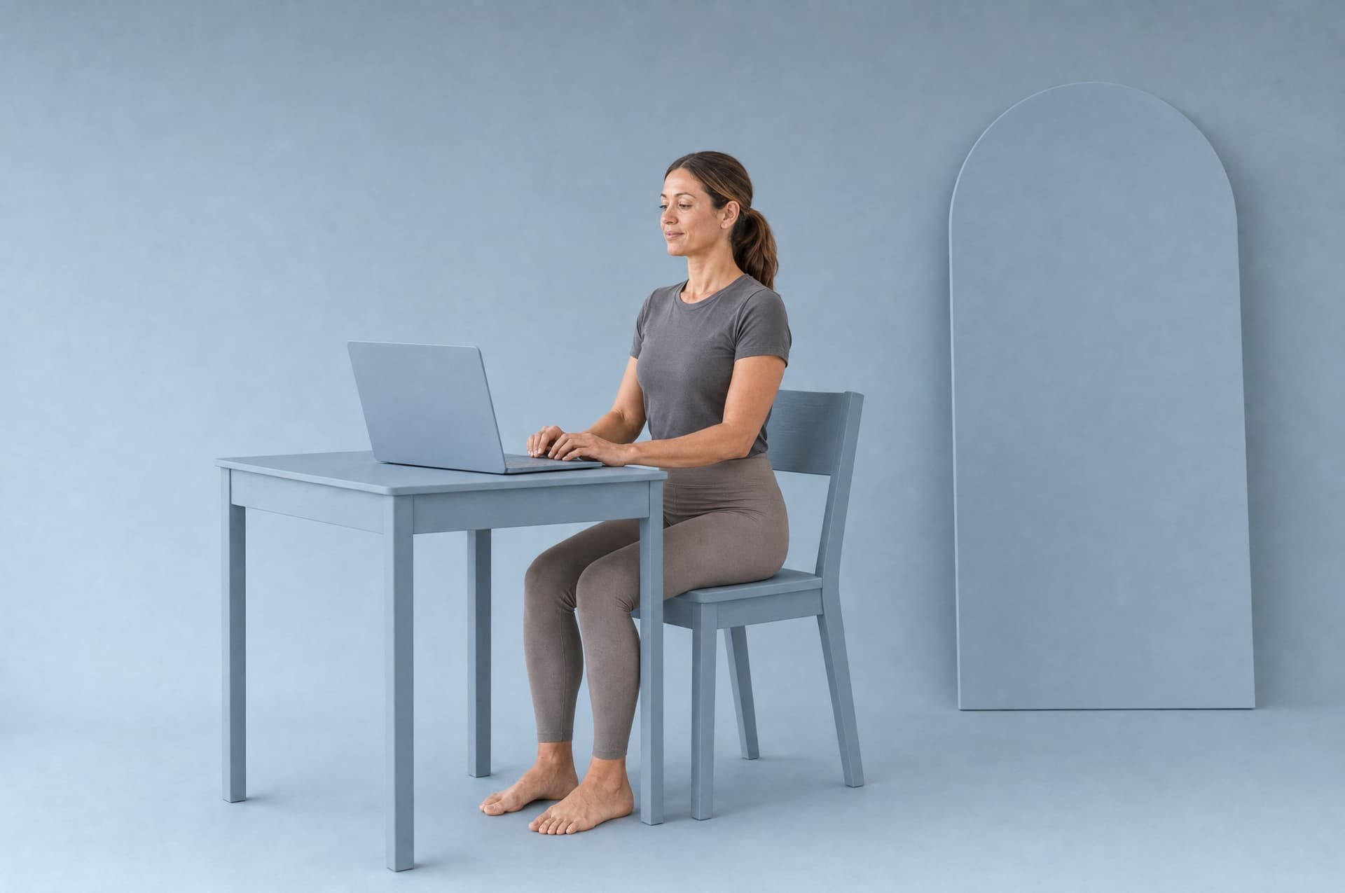A woman seated upright at a slate-blue desk in front of an open laptop, shoulders back, in a slate-blue studio.